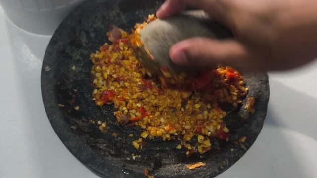 A person crushing ingredients on a black stone mortar to make an authentic and spicy Indonesian sambal.