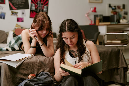Teenage girls studying together in bedroom, reading book and reviewing notes for homework, conveying focused learning and peer support. Useful for education, tutoring, exam prep
