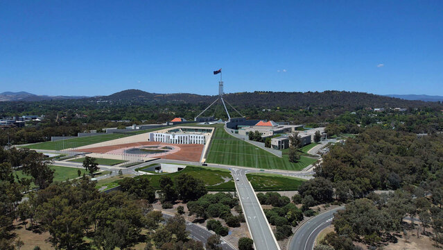 Aerial view of Parliament House with its iconic flagpole, expansive green lawns, and surrounding parklands in Canberra, Australian Capital Territory, Australia.