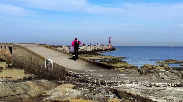 Long shot fixed camera video of an amateur fisherman strolling along the rocky shoreline of the R&iacute;o de la Plata during a sunny winter day in Montevideo, Uruguay, capturing natural landscape, leisure a