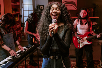Portrait of Black teenage girl singing into microphone while teen band playing keyboard, bass, guitar in rehearsal, representing youth music culture. Ideal for music education, teen talent promotion © AnnaStills