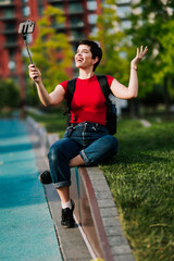 Obraz premium Smiling Caucasian female content creator recording a video with a smartphone mounted on a selfie stick while sitting outdoors in a city park