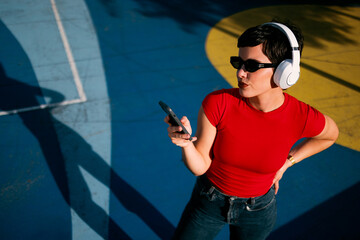 Fototapeta na wymiar Young Caucasian woman wearing wireless headphones and sunglasses while checking her smartphone in a colorful urban outdoor setting