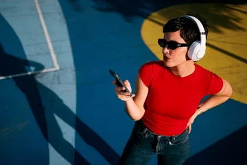 Fototapeten Pony Young Caucasian woman wearing wireless headphones and sunglasses while checking her smartphone in a colorful urban outdoor setting  © djile