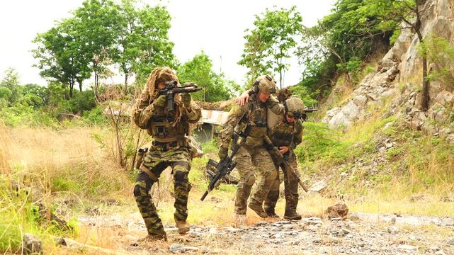 A group of soldiers in uniform, carrying guns, provide security, while another soldier assists the wounded during a training exercise while playing a BB gun game.
