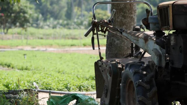 An old farm tractor stands by the edge of a green field near a tree, with countryside in the background. Traditional agriculture.