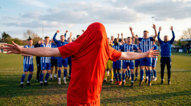 Amateur soccer player in red celebrating as his teammates cheer on a grass pitch