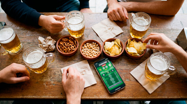 Overhead view of friends with beers snacks and a sports betting app on a smartphone