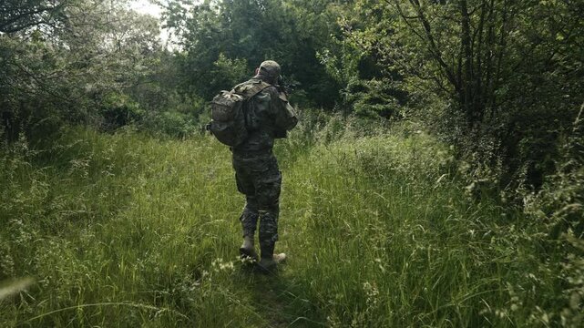 Camouflaged soldier traversing a footpath in the middle of rural wilderness. Carrying an AR-15 carbine rifle with a red collimator and a magnifier