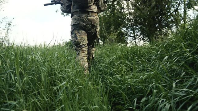 Camouflaged soldier traversing a footpath in the wilderness. Carrying an AR-15 carbine rifle with a red collimator and a magnifier