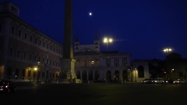 Lateran Obelisk, ancient Egyptian obelisk next to Basilica, glowing lanterns, city traffic and dark blue sky with gloowing moon - evening view in Rome in Italy. Topics: antiquity, tourist attraction