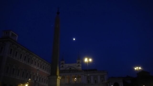 Lateran Obelisk, ancient Egyptian obelisk with Christian cross next to Basilica, glowing lanterns and dark blue sky with gloowing moon - evening view in Rome in Italy.