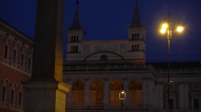 Lateran Obelisk, ancient Egyptian obelisk with Christian cross next to Basilica, glowing lantern and dark blue sky with gloowing moon - evening view in Rome in Italy.