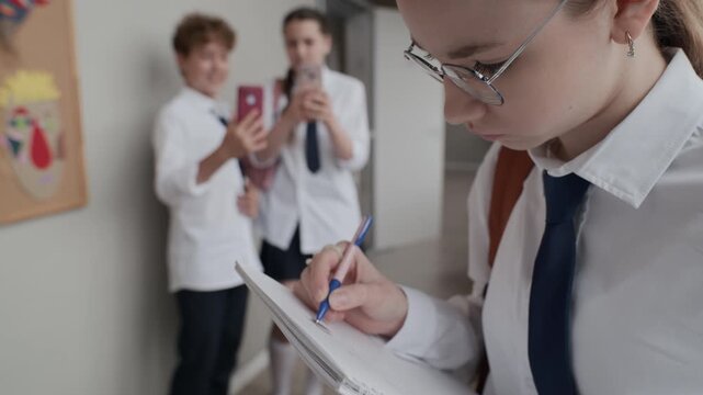 Caucasian teenage girl writing in notebook while classmates recording her on smartphones in school hallway. Perfect for cyberbullying awareness and school mental health campaigns