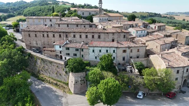 Veduta aerea di un borgo storico arroccato su una collina, con edifici in pietra, tetti in terracotta, mura antiche e campanile dominante, circondato da colline verdi e campi coltivati