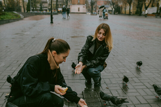 Female friends feeding pigeons on rainy autumn day in city square