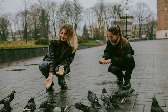 Female friends feeding pigeons on rainy autumn day in city square