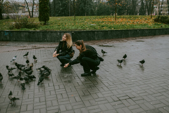 Female friends feeding pigeons on rainy autumn day in city square