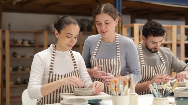 Young girl craftsperson in striped aprons busy molding clay products in lesson with female teacher. Amateur sculpting plate of raw clay under supervision of teacher