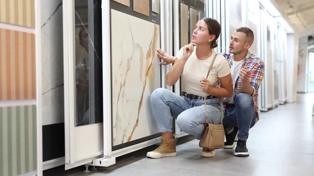 Positive couple, young woman and man, examining technical stone tile samples displayed on sliding exhibit stand in home improvement store, considering colors and textures for renovation project