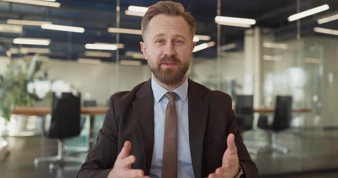 Businessman executive speaker in office on webcam. He addresses viewers during a video conference, hands on a desk in a modern boardroom. Strong corporate communication and leadership message.