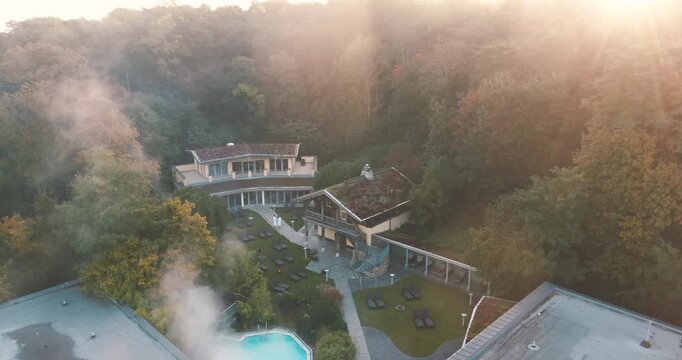 Aerial drone shot of a hotel complex surrounded by dense autumn forest with steam rising from a swimming pool