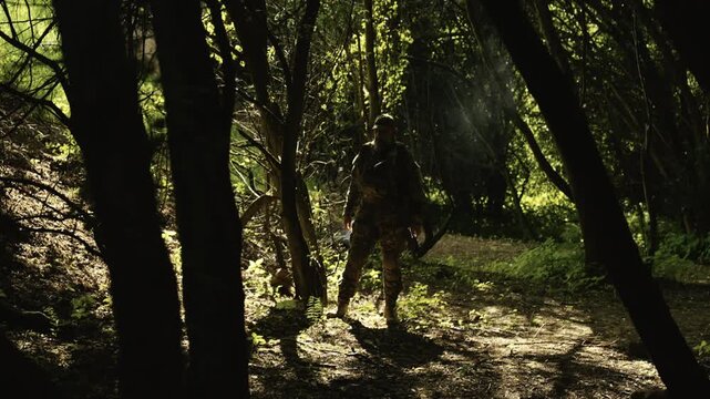 Camouflaged soldier in the middle of a dark forest on a recon mission. Smoking and holding a AR-15 carbine rifle with a red collimator and a magnifier