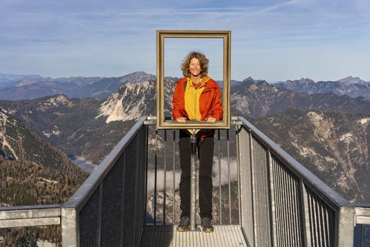 A hiker stands on the 5fingers viewpoint and looks through the picture frame. View from the Dachstein Krippenstein. Autumn, good weather. Some clouds in the mountains. Lake Hallstatt, Salzkammergut, Upper Austria, Austria
