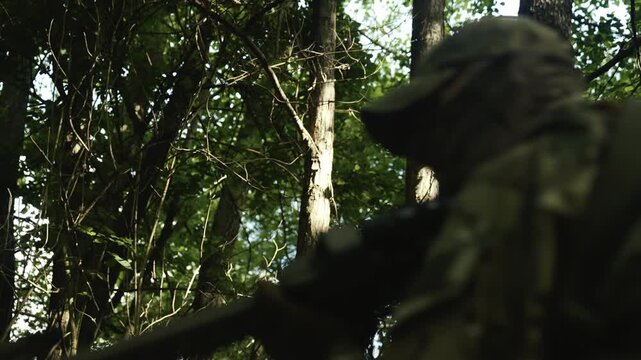 Camouflaged soldier in the middle of a dark forest on a recon mission. Reloading and aiming a AR-15 carbine rifle with a red collimator and a magnifier