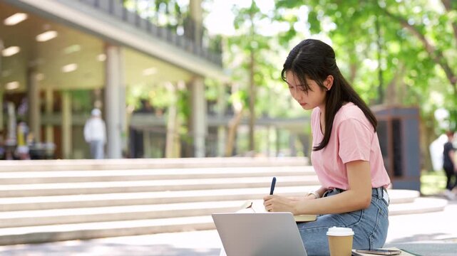 Focused asian teen girl student sitting outdoor serching on laptop writing notes in notebook distance learning studying online. college school education, e learning concept.