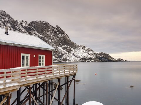 Traditional red wooden house on pegs by the fjord, snow-capped mountains behind, Nusfjord, Flakstad&oslash;ya, Lofoten, Norway