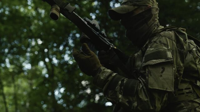 Camouflaged soldier in the middle of a forest on a recon mission. Reloading and aiming a AR-15 carbine rifle with a red collimator and a magnifier