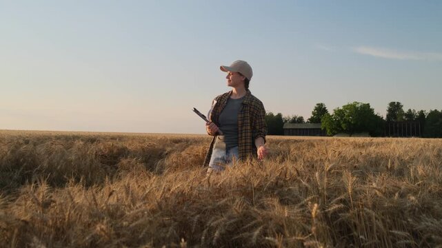 A young farmer woman in a cap and a plaid shirt stands in a field where ripe rye grows. She looks at her good harvest and smiles. The theme of the autumn harvest.