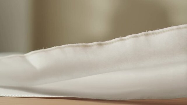 A hand gently lifts and smooths a soft, white fabric on a table, highlighting its delicate texture and fine craftsmanship under warm, natural lighting.
