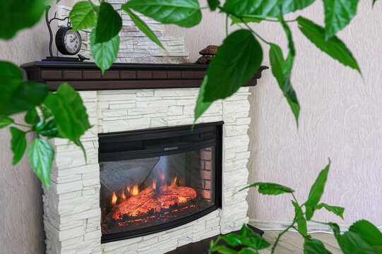 modern electric fireplace features a white stone-veneer surround, a dark wooden mantel with decor, a realistic flame effect, and is framed by a green plant and light wood flooring