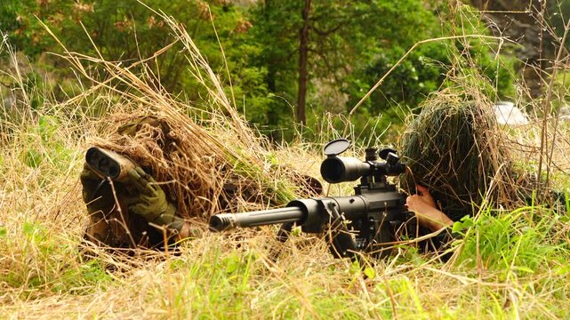  Two soldiers in camouflage uniforms stand in a field, with one soldier randomly aiming his rifle and another providing coordinates to a spotter to monitor the target.
