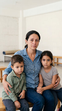 Madre e hijos con rostros de preocupaci&oacute;n y tristeza sentados en un refugio, reflejando el impacto emocional del trauma familiar, la migraci&oacute;n y la vulnerabilidad social.
