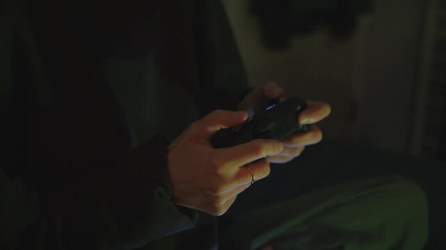 Gaming lifestyle, relaxation after work. Close-up of hands holding a wireless game controller while playing video games in a dimly lit, cozy room.
