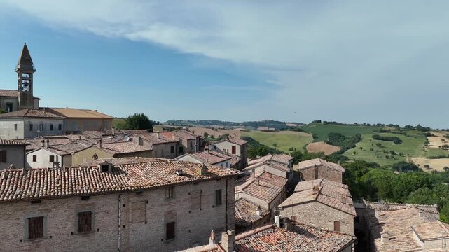 Veduta aerea di un piccolo borgo storico su collina, con edifici in pietra dai tetti in terracotta, strade strette e campanile, circondato da campi verdi e colline rurali