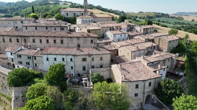 Veduta aerea di un piccolo borgo storico su collina, con edifici in pietra dai tetti in terracotta, strade strette e campanile, circondato da campi verdi e colline rurali