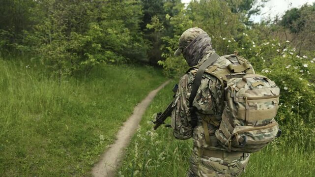 Camouflaged soldier in the middle of a field of grass on a recon mission. Walking and aiming a AR-15 carbine rifle with a red laser collimator and a magnifier