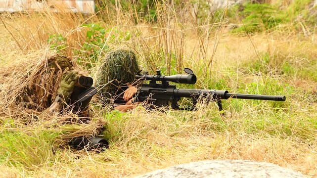 Two soldiers in camouflage uniforms stand in a field, with one soldier randomly aiming his rifle and another providing coordinates to a spotter to monitor the target.
