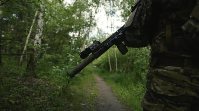 Legs of a camouflaged soldier in the middle of a footpath in a forest on a mission. Walking and aiming a AR-15 carbine rifle with a red laser collimator and a magnifier