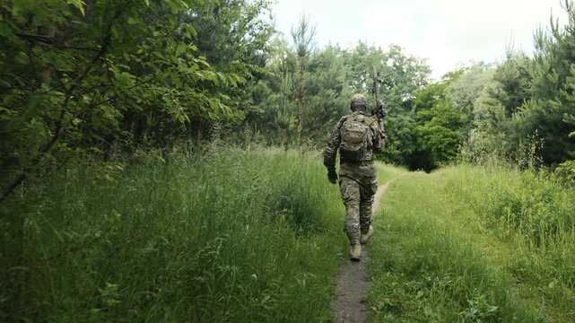 Legs of a camouflaged soldier in the middle of a footpath in a field of grass on a recon mission. Walking and aiming a AR-15 carbine rifle with a red laser collimator and a magnifier