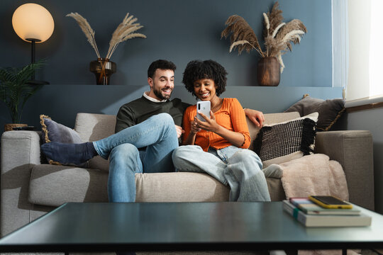 Young multi ethnic couple sitting on sofa using mobile phone app to share social media content at home