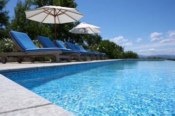 Fototapeta premium Luxurious infinity pool with sparkling blue water and empty lounge chairs under white umbrellas on a sunny day.