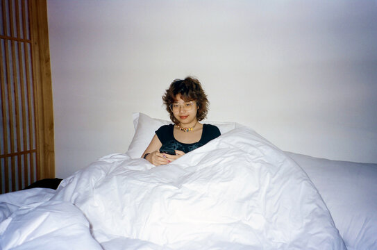 Young Woman Sitting Up in Bed With Phone and White Duvet