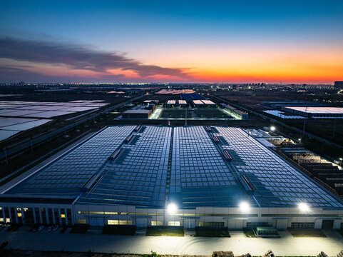 Aerial view of large industrial factory complex at dusk with illuminated glass roofs and urban skyline in background