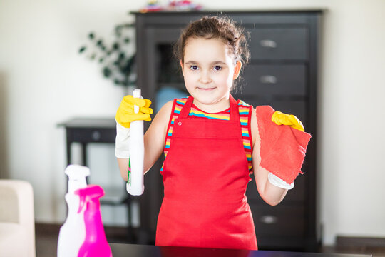 A child in a red apron and yellow gloves cleaning a table with a spray bottle and cloth