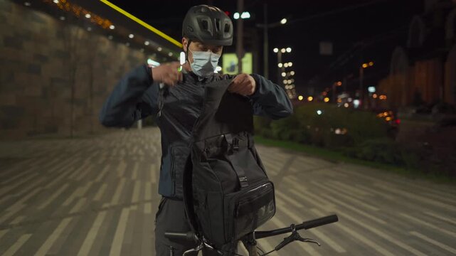 Urban cycling video showing a man with bicycle helmet and protective face mask organizing his backpack after a ride. Night city background highlights sustainable transport and commuter lifestyle.